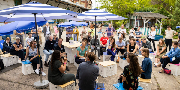 Menschen sitzen auf weißen Kisten im Halbkreis um 3 Personen, die offenbar im mittelpunkt der Unterhaltung stehen.